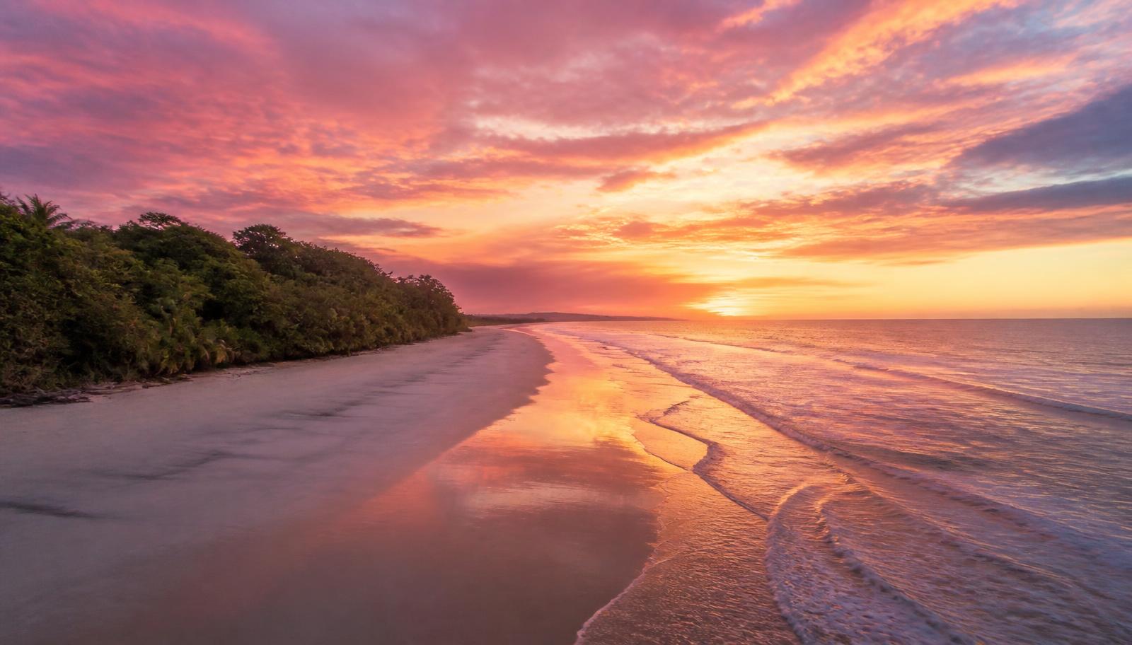 Playa larga y vacía de arena blanca en Costa Rica al amanecer con un cielo rosado y dorado reflejado en la arena mojada
